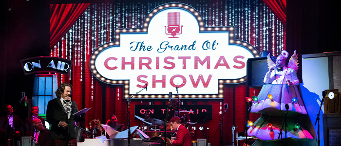 A man stands at a microphone on the left, in the center sits a pianist, and to the right is a man dressed as a christmas tree / angel