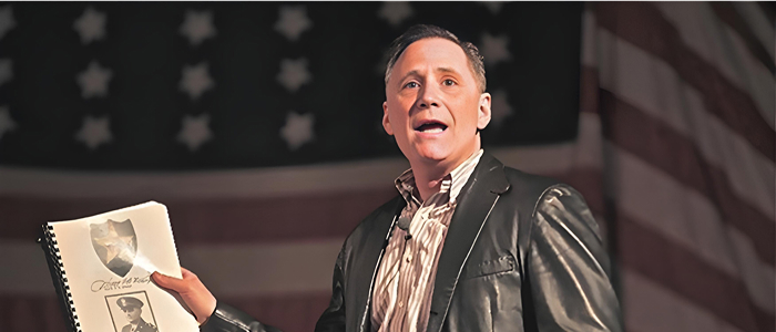 A man holding a book in his right hand while speaking on stage in front of American flag backdrop