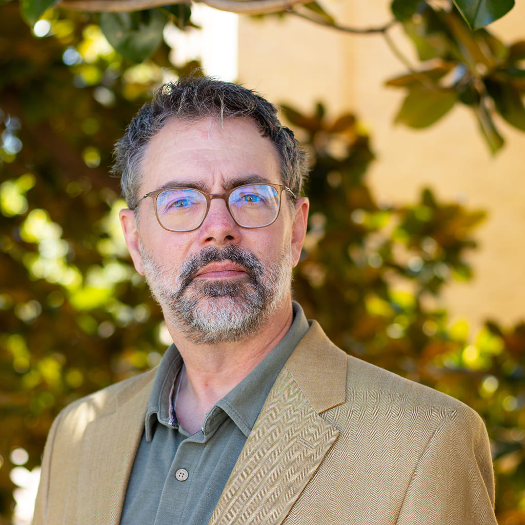 A man standing in front of a tree in a suit and with glasses