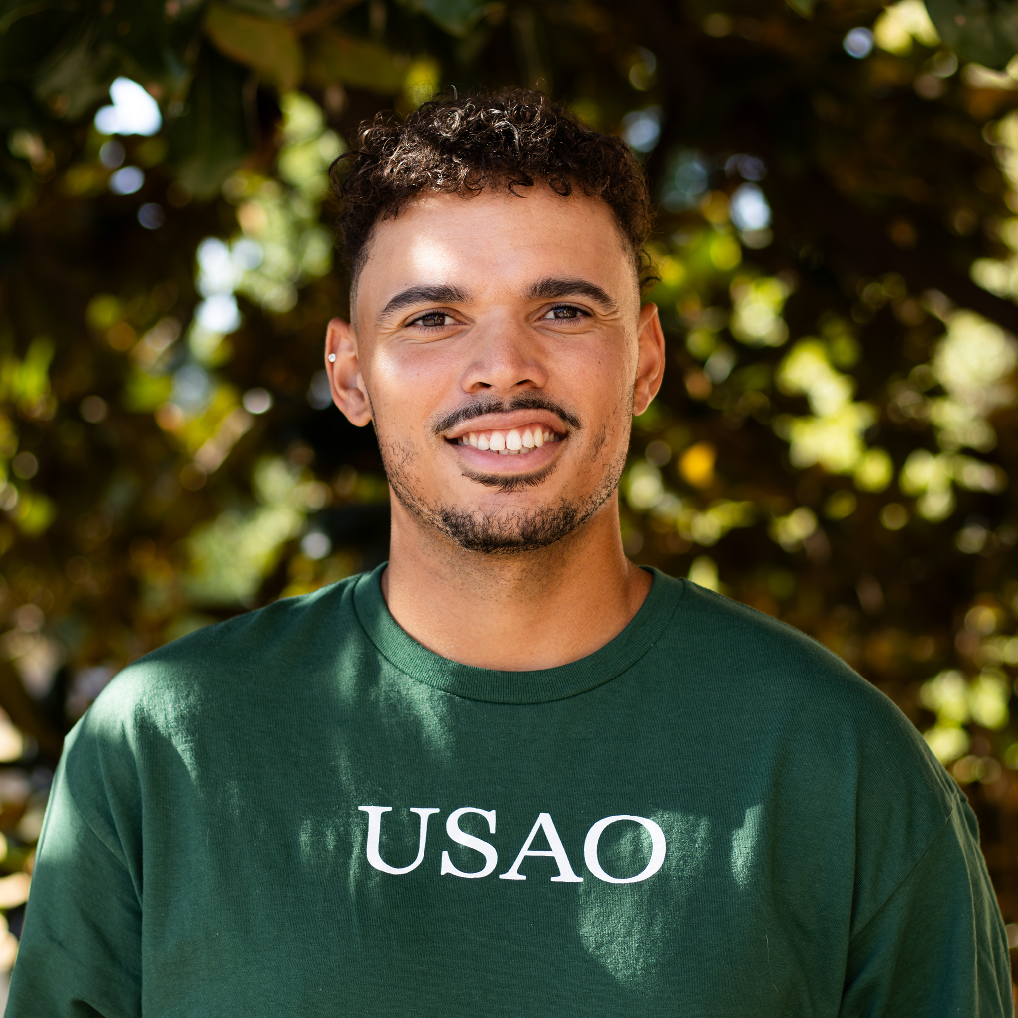 A man in a green t-shirt standing and smiling in front of a tree
